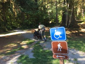 Wheelchair hiking sign in foreground, person pushing a wheelchair along gravel path in background.